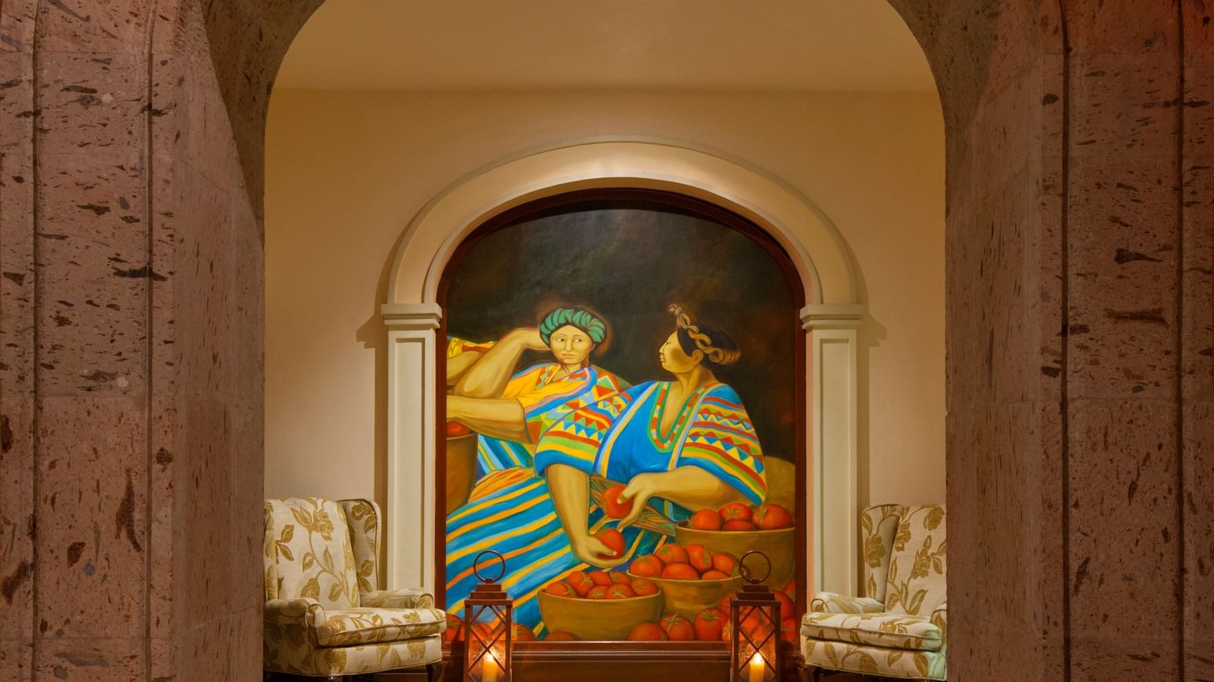 Colorido mural de mujeres con fruta bajo un arco, con dos sillas y faroles en Tomates en Hacienda Del Mar Los Cabos.