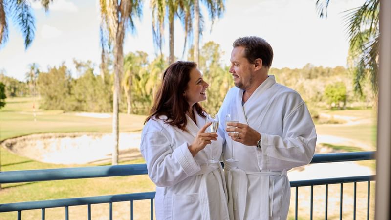 Couple in bathrobes clinking wine glasses on a balcony overlooking a golf course.