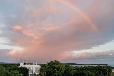 Pink sunset sky with rainbow and white hotel building among lush trees at El Blok