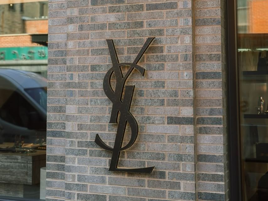 YSL logo on brick wall next to glass storefront with reflection of van and buildings.