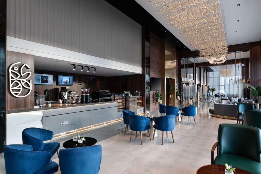 Blue tub chairs by a modern coffee counter under a tall wood ceiling in Khva Cafe at Warwick Al Taif Hotel
