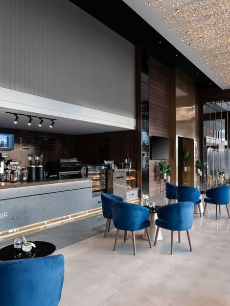 Blue tub chairs by a modern coffee counter under a tall wood ceiling in Khva Cafe at Warwick Al Taif Hotel