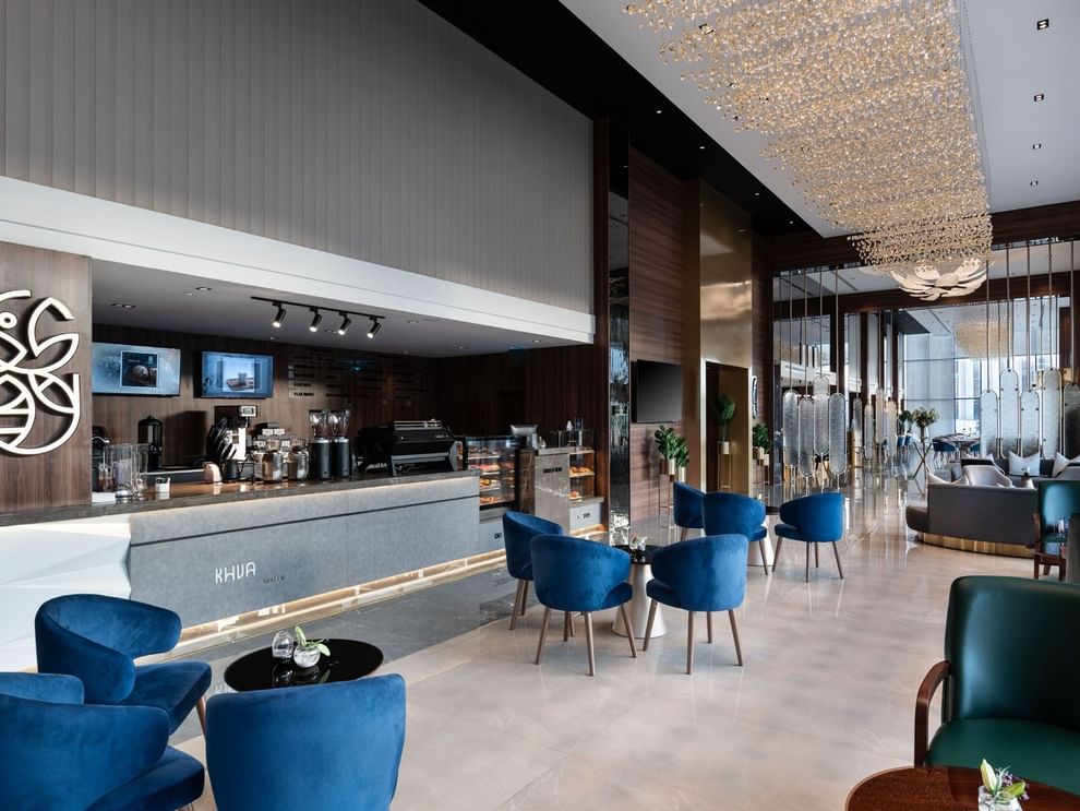 Blue tub chairs by a modern coffee counter under a tall wood ceiling in Khva Cafe at Warwick Al Taif Hotel