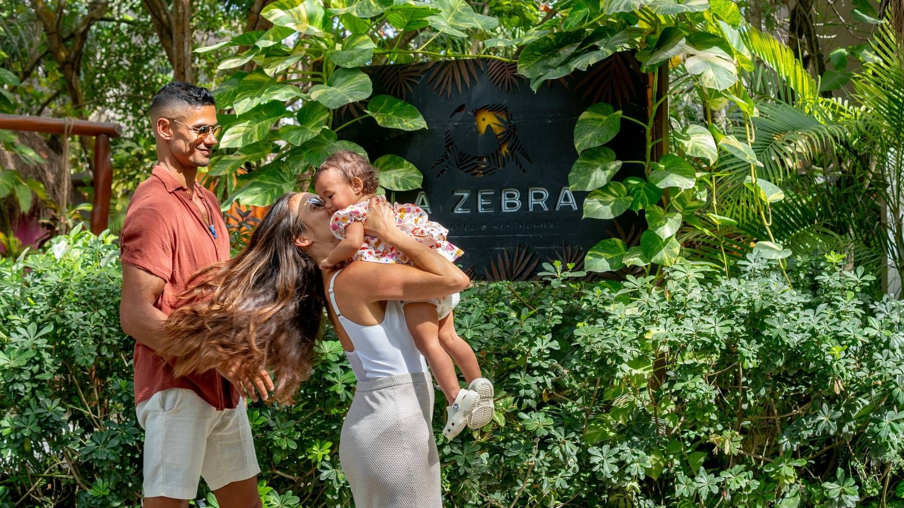 Parents cuddling their child beside La Zebra's sign and tropical greenery