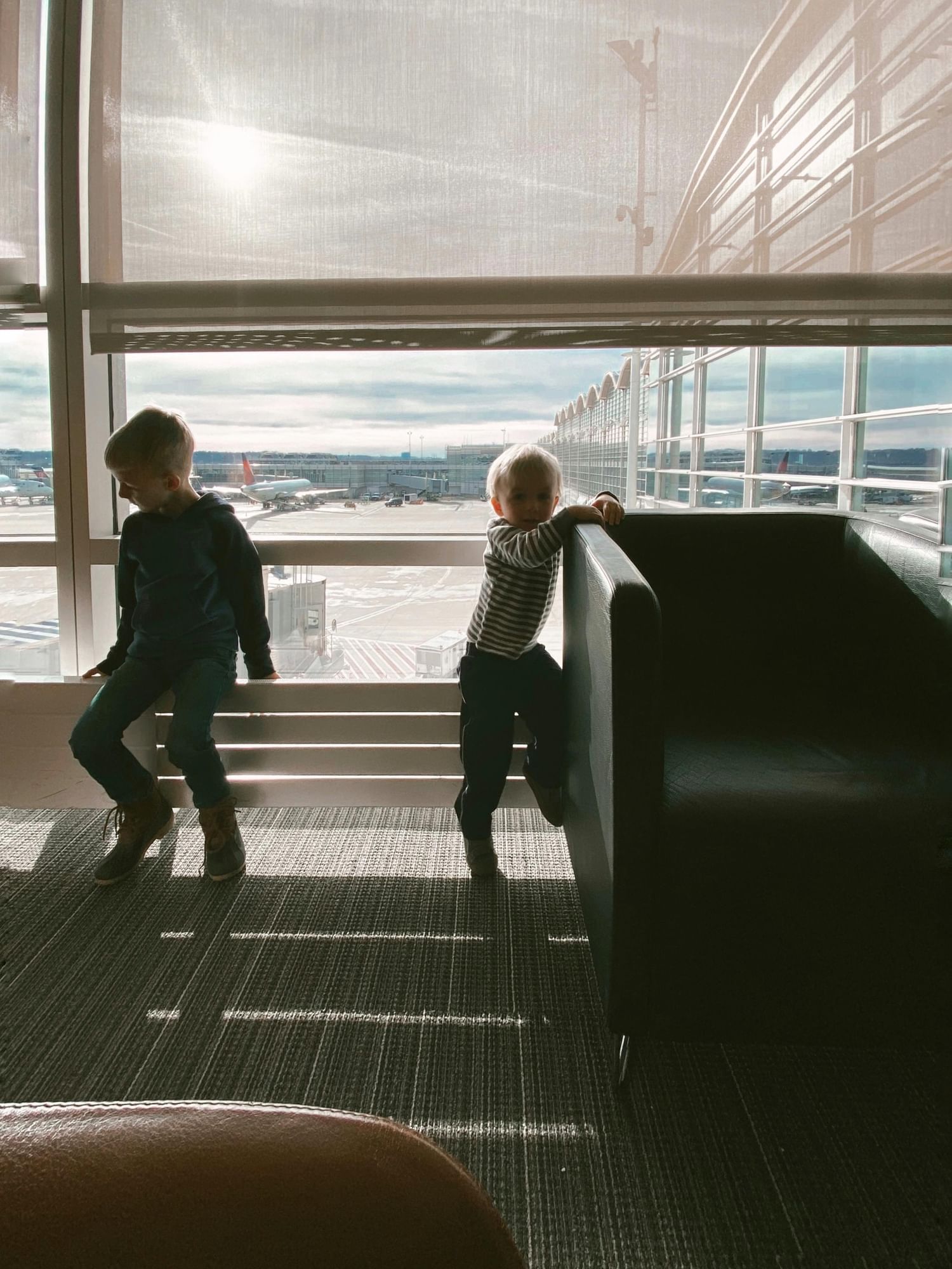 Two children at an airport gate silhouetted in front of a window overlooking an airport loading dock. 