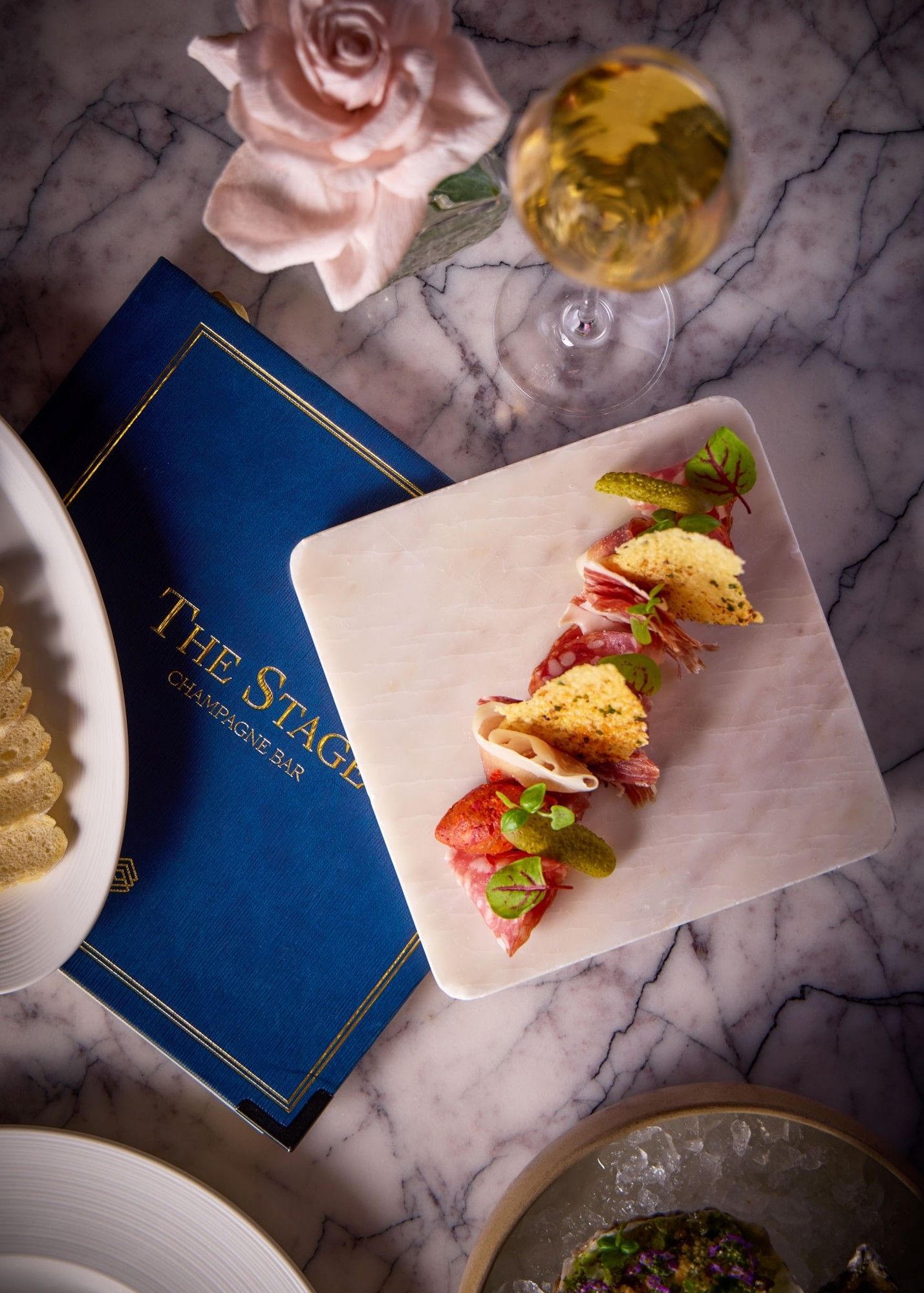 Gourmet appetizer with cured meats and crispy garnish beside Champagne on marble table at The Stage in The Londoner Hotel 