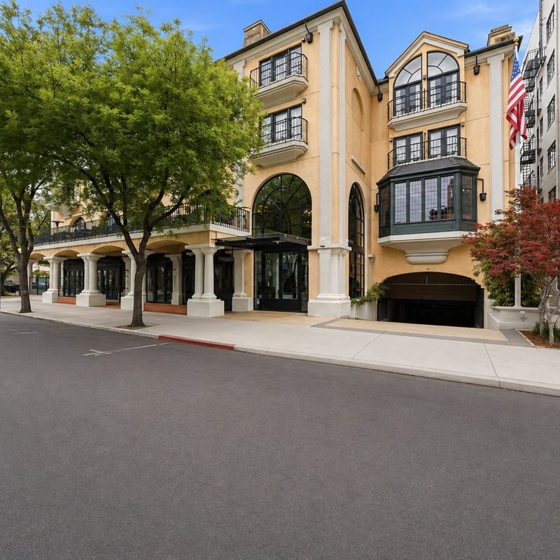 Street view of a Mediterranean-style hotel building with arched entrance and tree-lined sidewalk at el PRADO Hotel