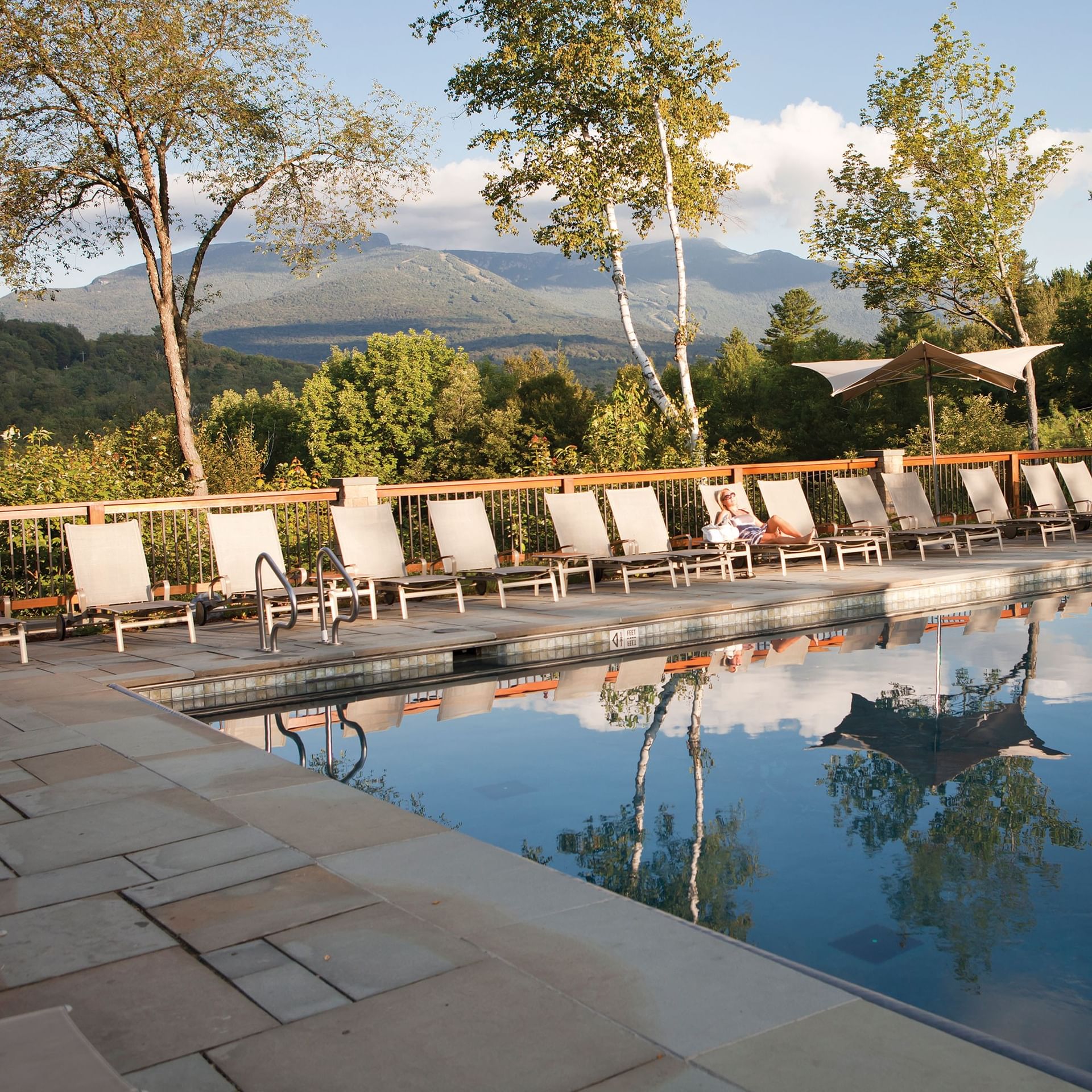 Woman relaxing on a lounge chair beside a pool with mountains in the background.