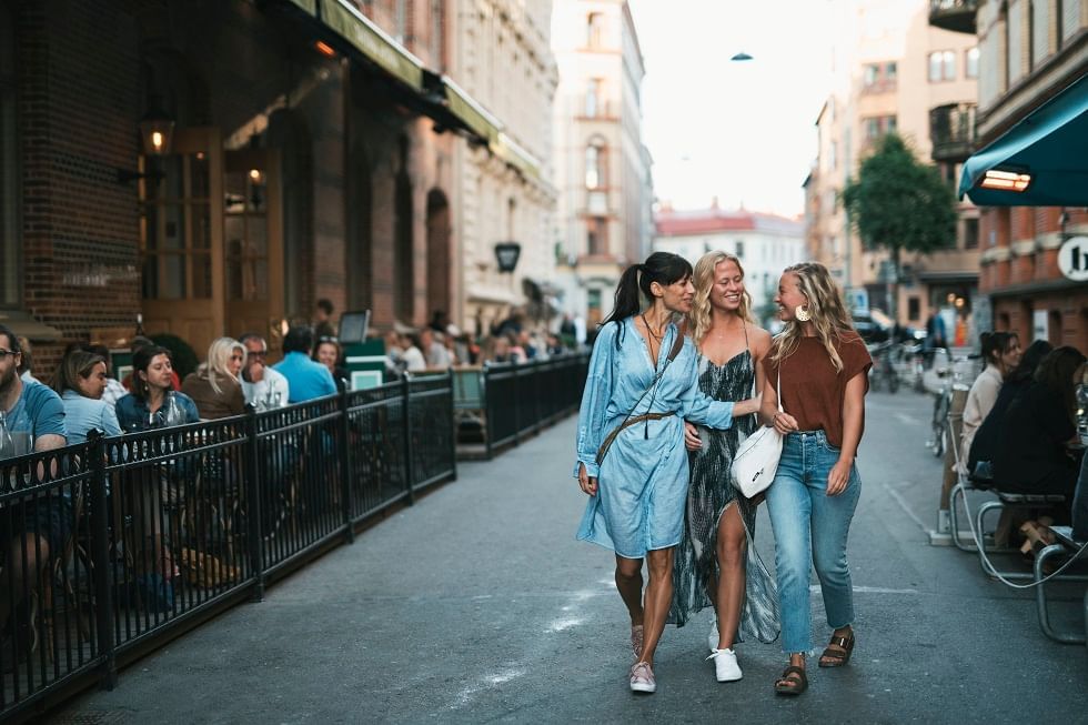 Three women walking together on a city street, surrounded by outdoor diners and tall buildings.