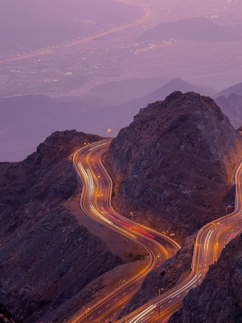 Winding mountain road with light trails by rocky peaks under a purple dusk sky near Warwick Al Taif Hotel