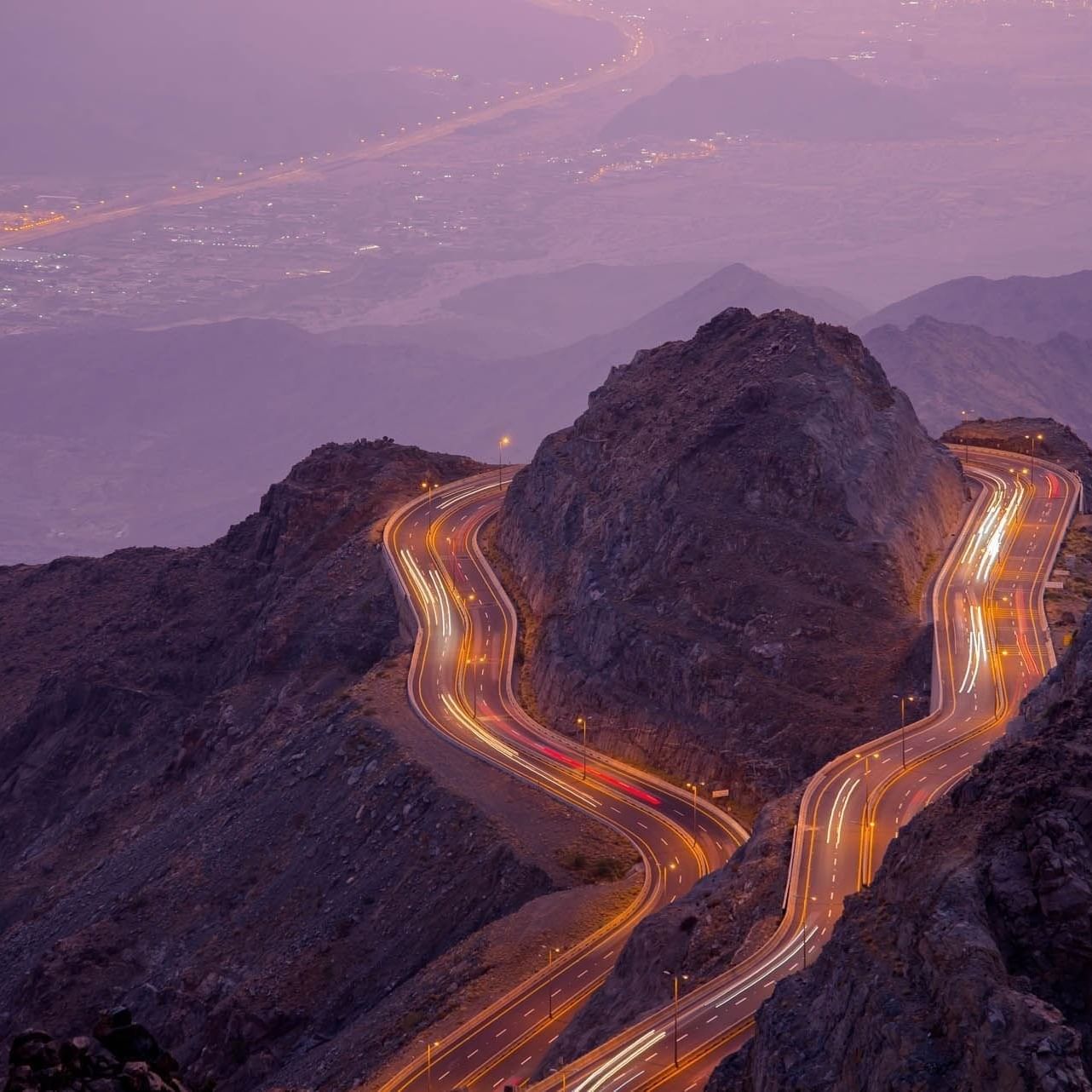 Winding mountain road with light trails by rocky peaks under a purple dusk sky near Warwick Al Taif Hotel