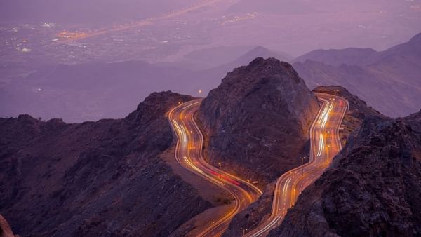 Winding mountain road with light trails by rocky peaks under a purple dusk sky near Warwick Al Taif Hotel