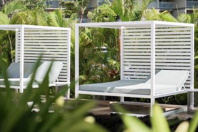 White outdoor cabana beds with pillows and lounge chairs, surrounded by lush green tropical plants at the Maui Coast Hotel