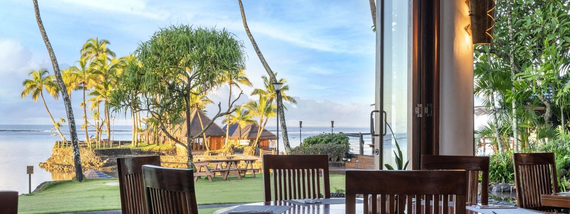 Dining table with wooden chairs by an open glass door under a warm lamp in Bula Brasserie at Warwick Fiji Resort and Spa