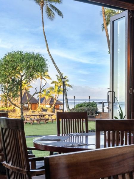 Dining table with wooden chairs by an open glass door under a warm lamp in Bula Brasserie at Warwick Fiji Resort and Spa