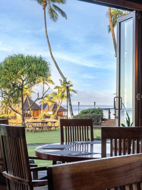 Dining table with wooden chairs by an open glass door under a warm lamp in Bula Brasserie at Warwick Fiji Resort and Spa