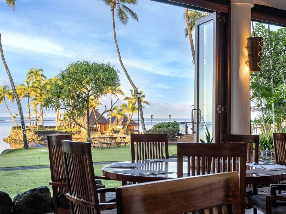Dining table with wooden chairs by an open glass door under a warm lamp in Bula Brasserie at Warwick Fiji Resort and Spa