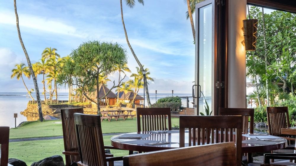 Dining table with wooden chairs by an open glass door under a warm lamp in Bula Brasserie at Warwick Fiji Resort and Spa