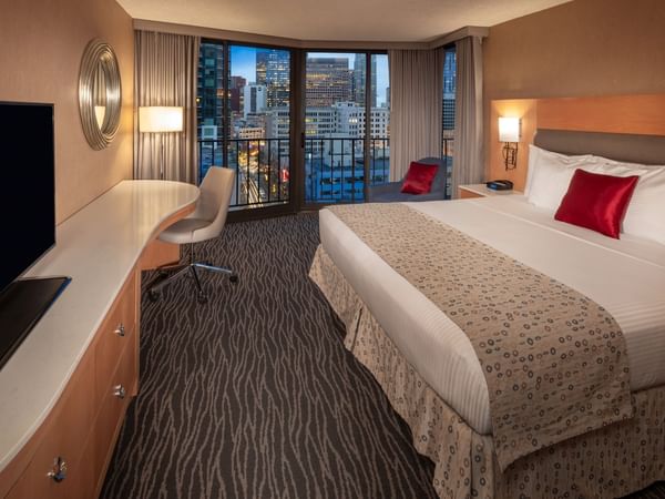 A hotel room with a large bed, desk with a TV, chair, and balcony overlooking a cityscape at night.