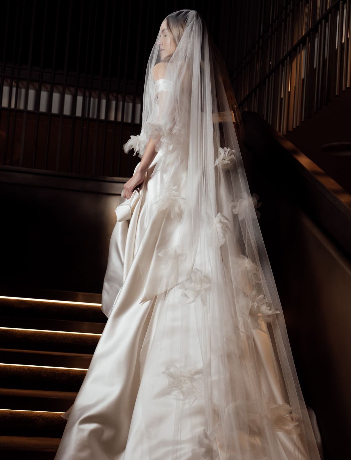 A bride in a long, flowing white wedding gown and veil ascends a dark, grand staircase at The Londoner Hotel