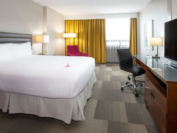 A hotel room with a bed, desk, chair, TV, and yellow curtains over a window.