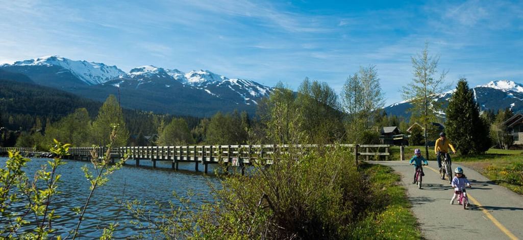 Family biking along the Valley Trail in Whistler with snow-capped mountains and a wooden bridge in the background.