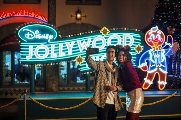 A couple takes a selfie in front of a Disney Jollywood Nights neon sign with a waving gingerbread character.