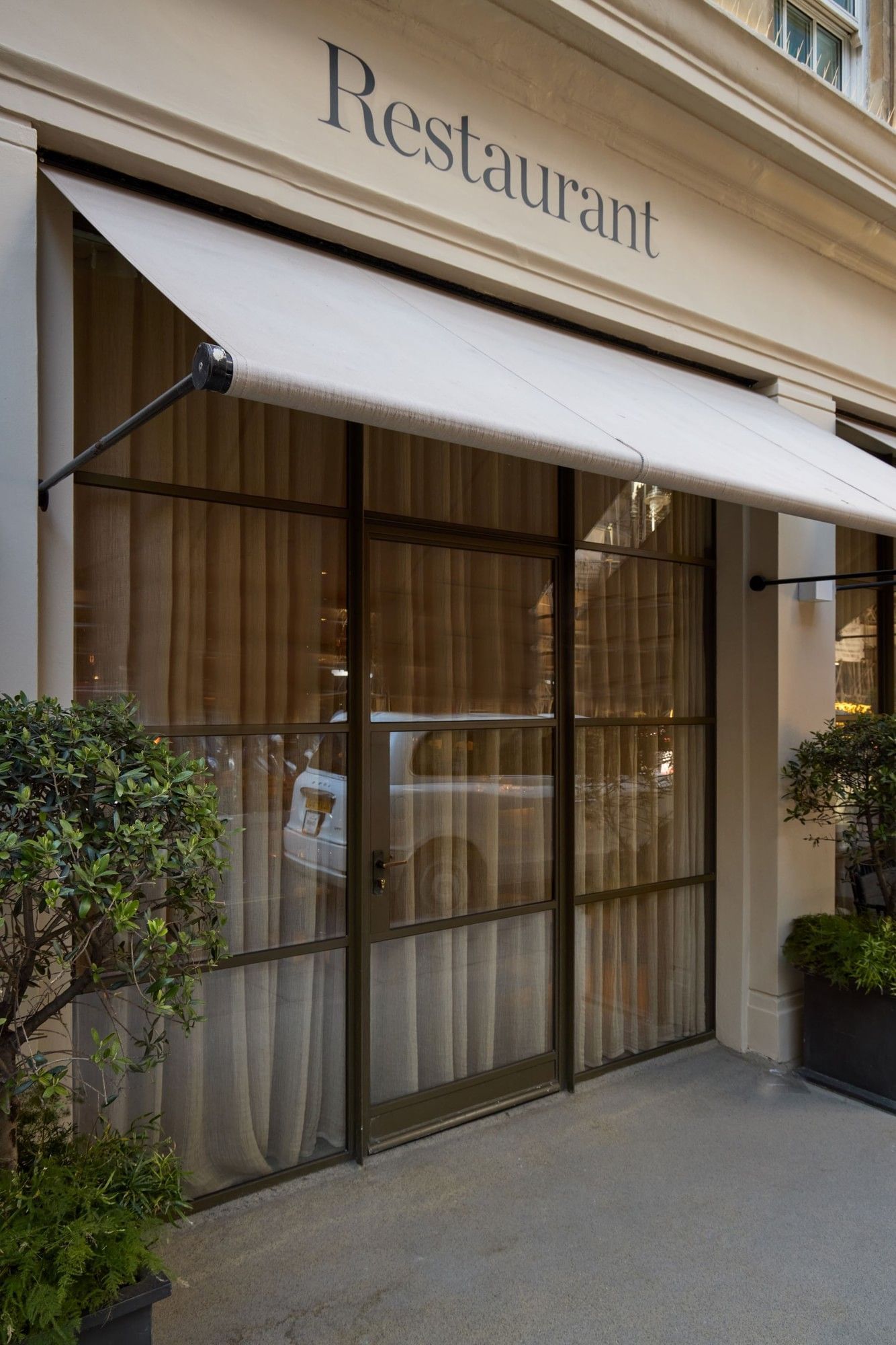 May Fair Kitchen restaurant entrance with white awning, glass doors, and green plants in planters.