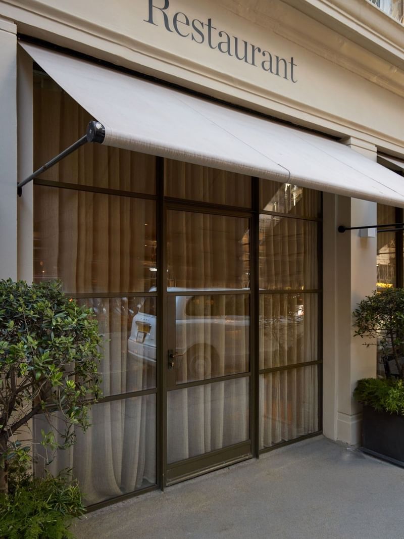 May Fair Kitchen restaurant entrance with white awning, glass doors, and green plants in planters.