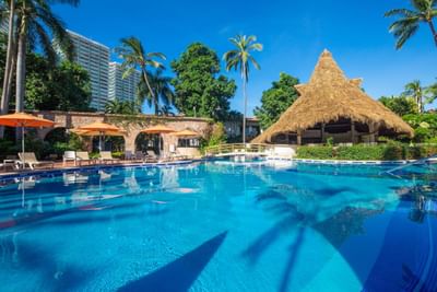 Resort pool with lounge chairs, umbrellas, and palm trees under a clear blue sky.
