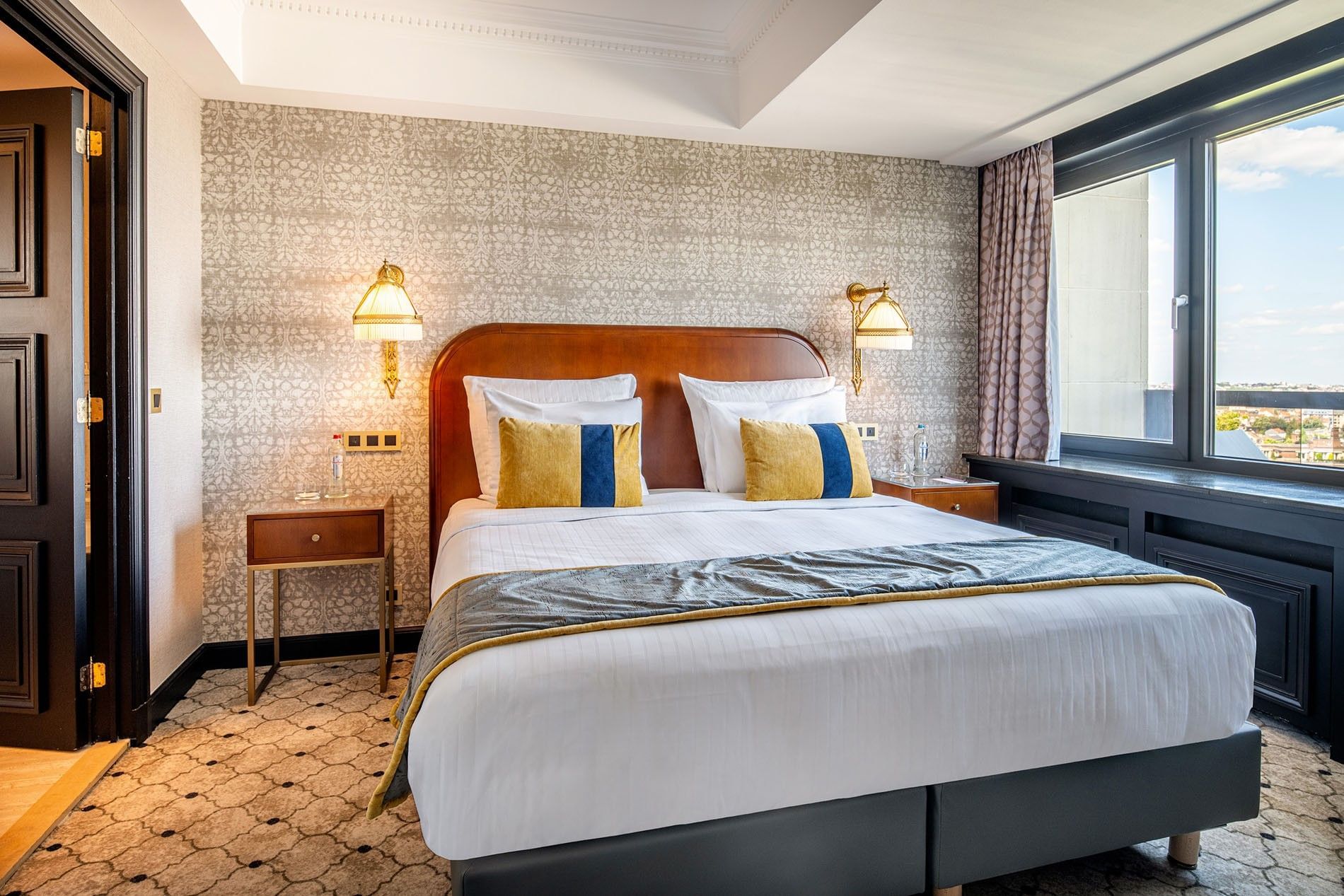 Plush bed between gold lamps with yellow cushions by a large window in Warwick Suite with terrace at Hotel Barsey by Warwick