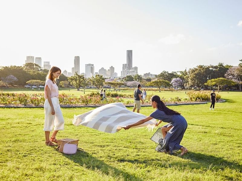 People setting up a picnic blanket on green lawn with city skyline in background at New Farm park