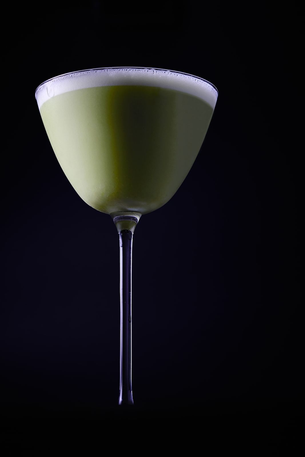 A martini glass holding a vibrant green cocktail with a foamy top against a dark background, a drink from The Londoner Hotel