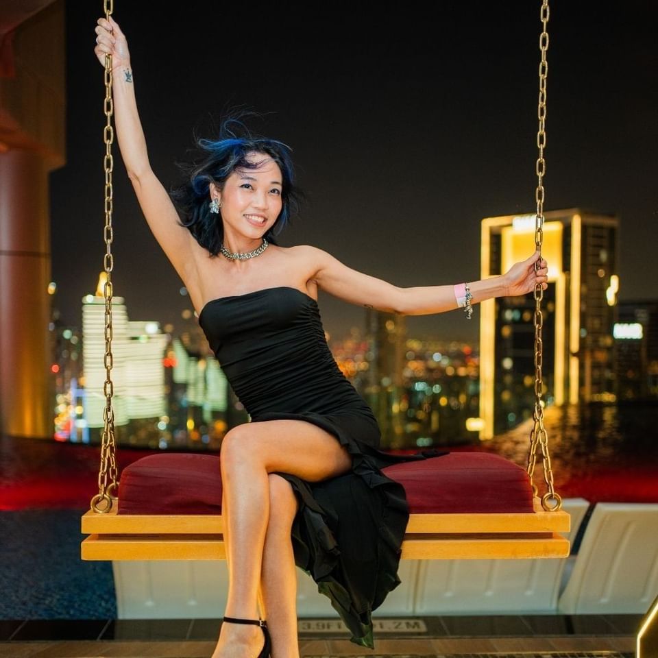 Smiling woman in a black dress posing on a wooden swing suspended over a rooftop pool at night inside Malibu Sky Lounge & Bar