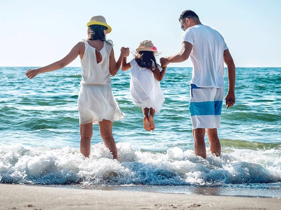 Familia de tres saltando juntos en la orilla de la playa con sombreros y ropa blanca bajo el sol brillante.