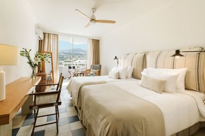 Two beds, chairs, desk with flowers, ceiling fan, and balcony with mountain view in a hotel room.