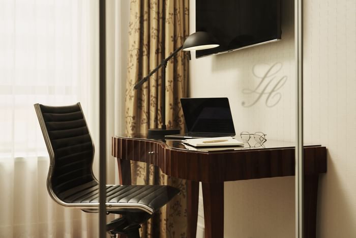 Desk with laptop, pen, and glasses, by black chair and curtained window, under wall-mounted TV and lamp.