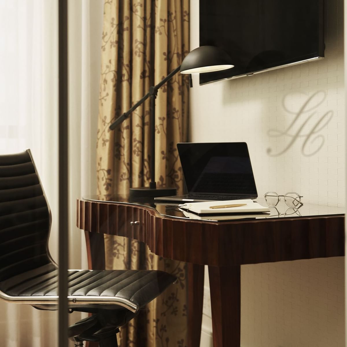 Office setup with a desk, laptop, lamp, chair, and TV in a room with curtains and a glass door.