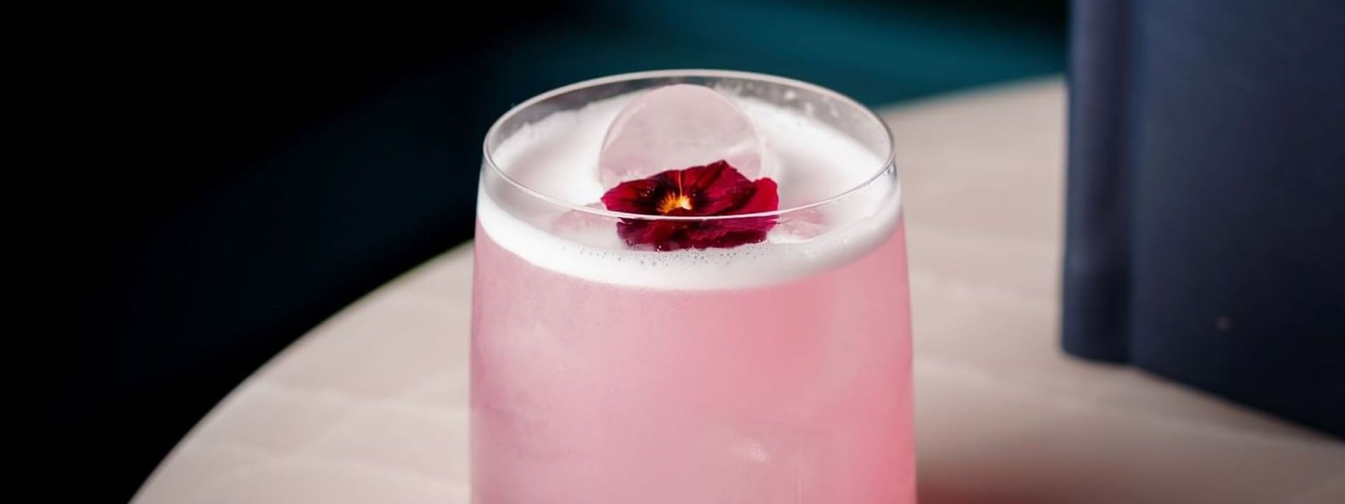 Pink cocktail with a foamy top and red edible flower garnish served on a round white table in Tom Brown at The Capital
