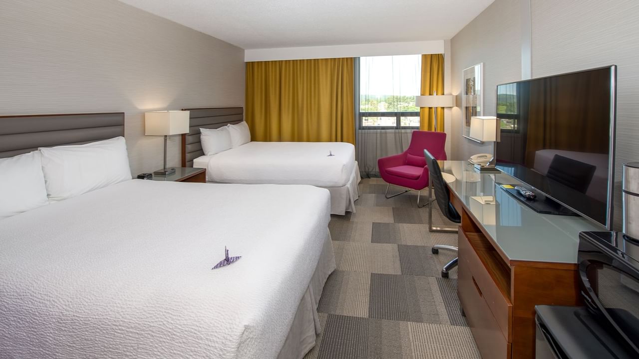 Two queen beds, large TV, armchair, and window with yellow curtains in a hotel room.