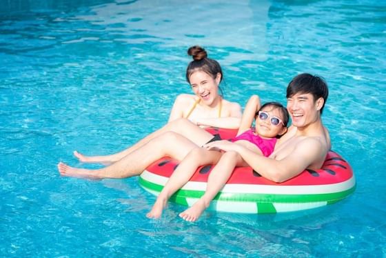 Family of three relaxing on a watermelon-shaped inflatable float in a sparkling blue pool at Rosen Inn at Pointe Orlando