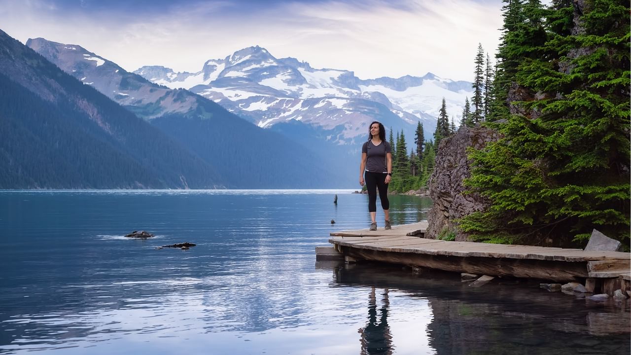 Woman standing on a wooden boardwalk by a serene lake with mountain backdrop.
