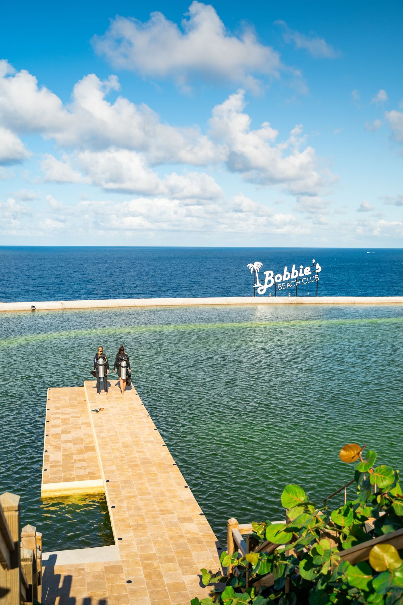 Twee duikers bereiden zich voor om het water in te gaan bij Bobbie's Beach Club met een zonnige hemel in Golden Rock Resort