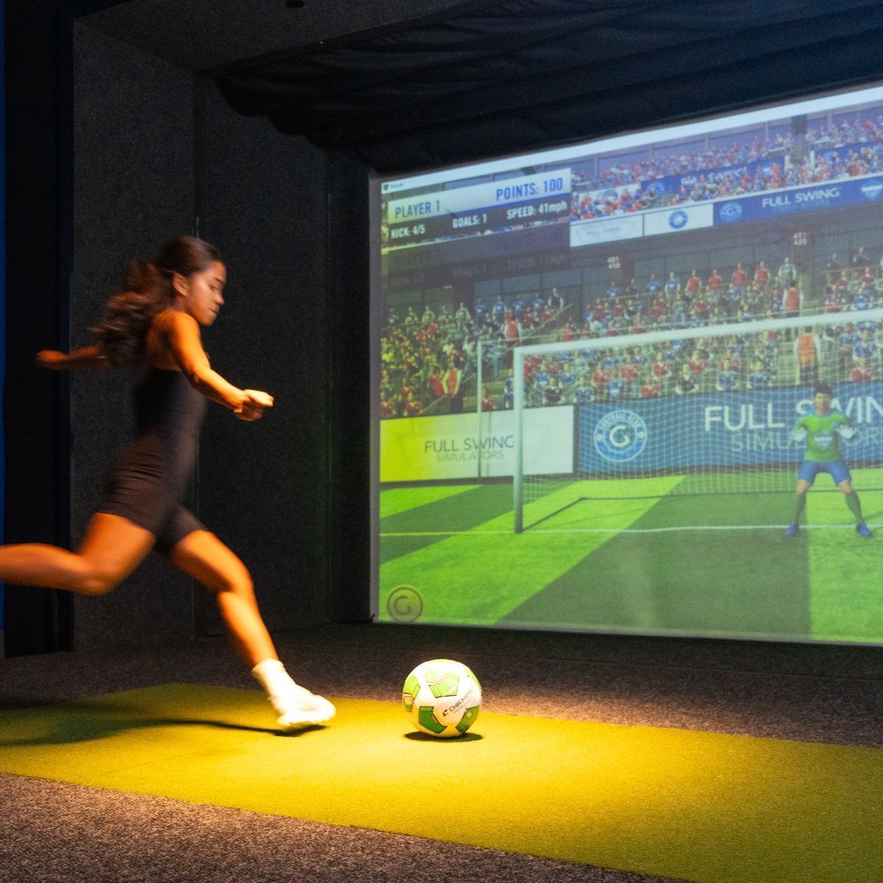 Woman in black sportswear playing soccer on a virtual reality simulator with a large screen showing a soccer game.