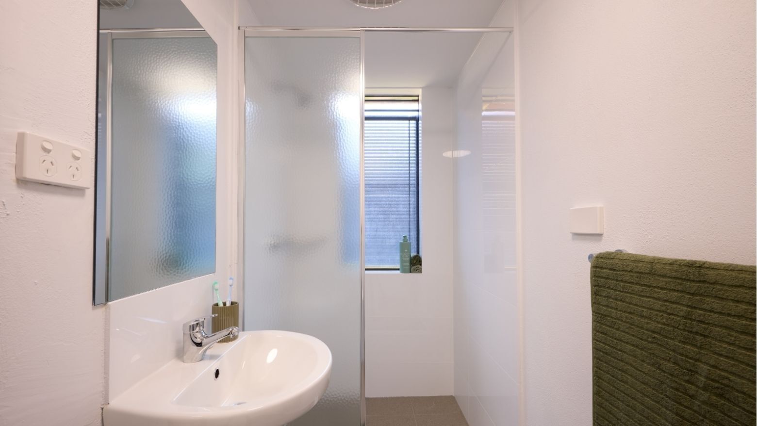 White bathroom with sink, mirror, glass shower enclosure, and green towel at UniLodge at Curtin University - Erica Underwood House.