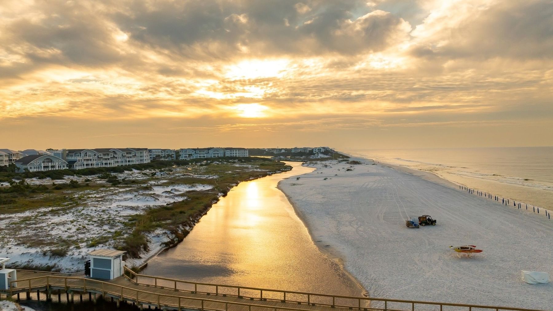 Sunset over a serene beach with a wooden bridge and distant buildings, highlighting Extended Stay Savings at Watersound Inn.