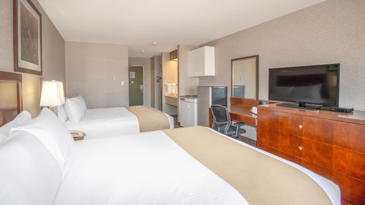 Two beds, desk with chair, TV, and refrigerator in a hotel room.