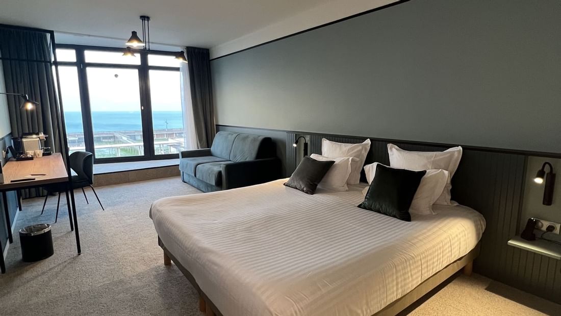 Chambre d'hôtel avec grand lit, canapé, bureau et vue sur la mer par la grande fenêtre.