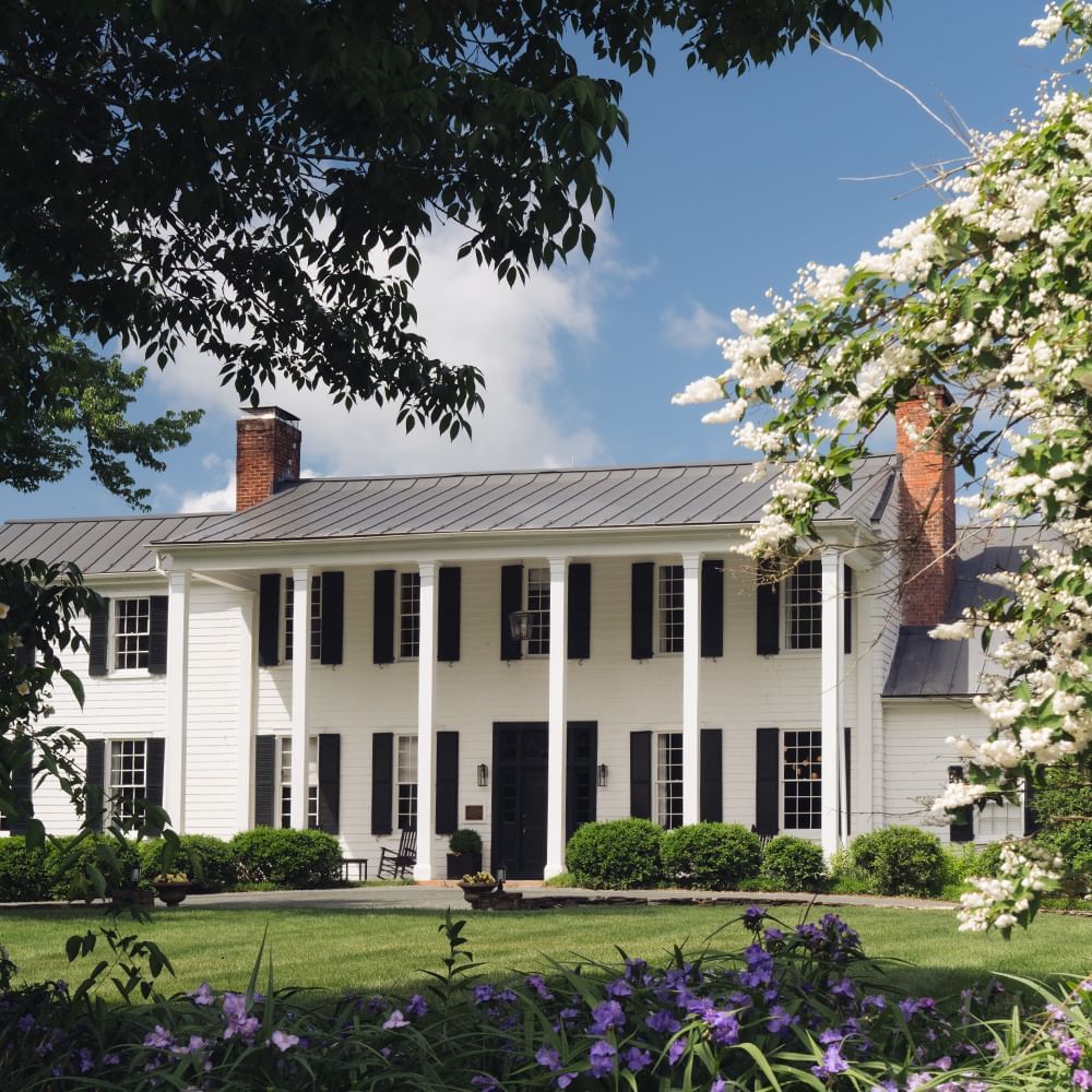 Facade of The Clifton Hotel in Charlottesville, VA, with black shutters, two chimneys, and a lawn with purple flowers.