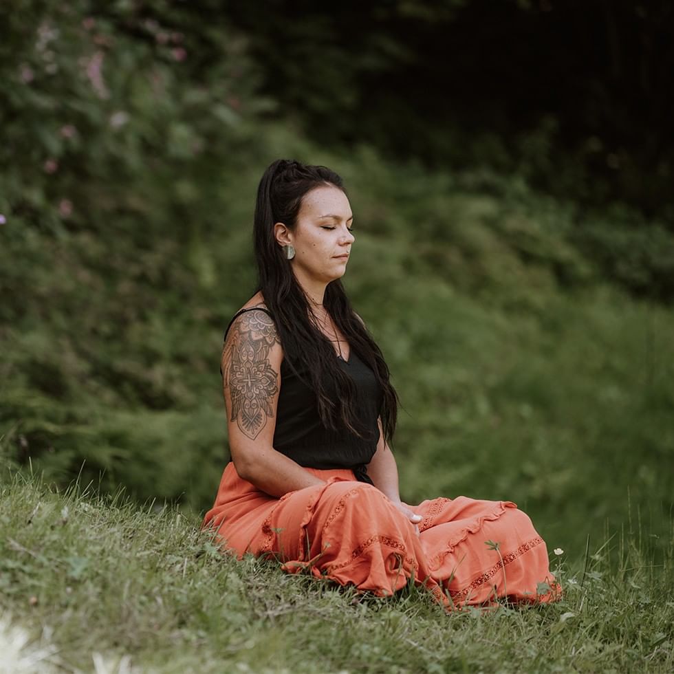 Frau mit Tattoos meditiert im Schneidersitz auf einer Wiese in der Natur.